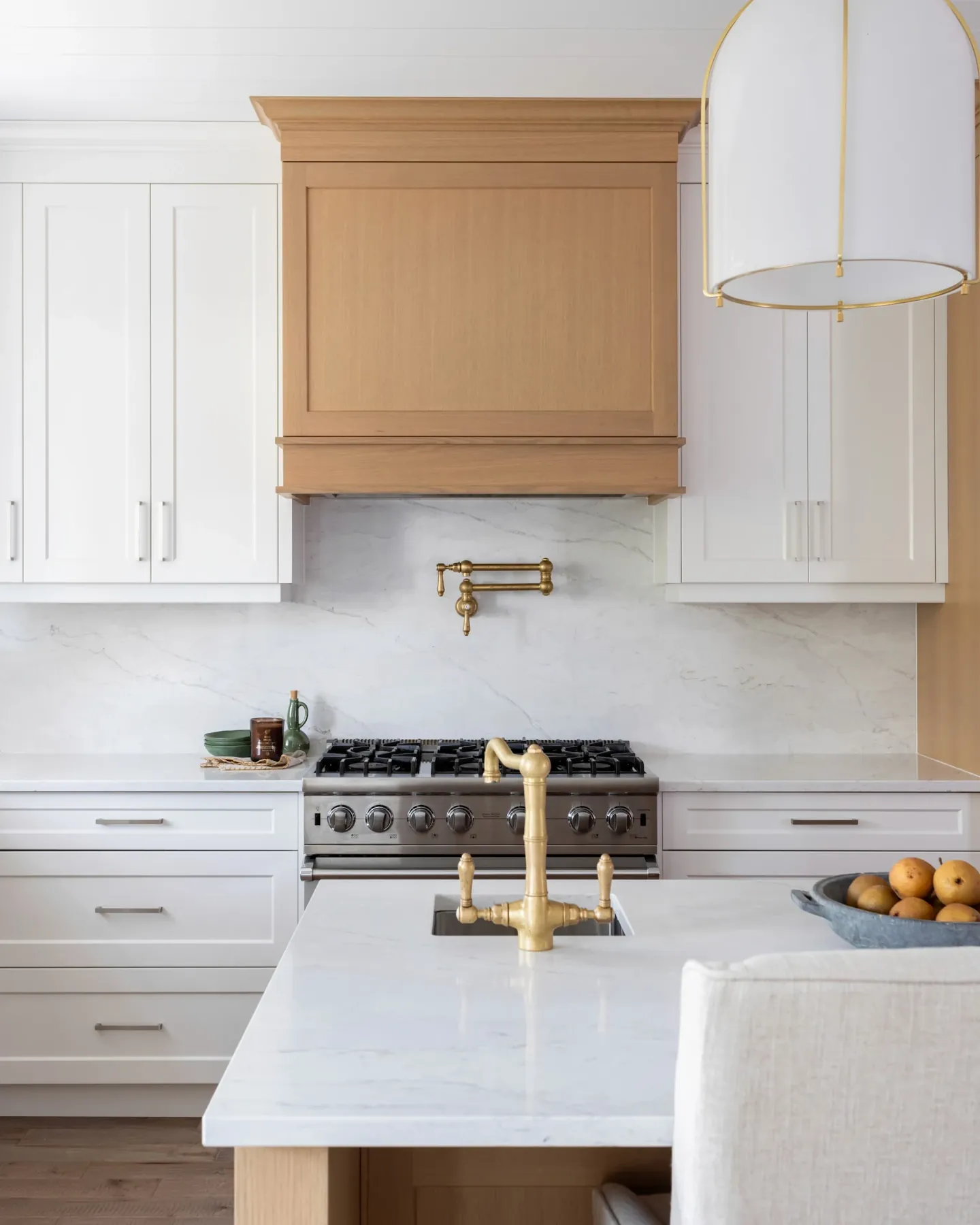 Gas range beneath a white oak hood with a brass pot filler and marble backsplash